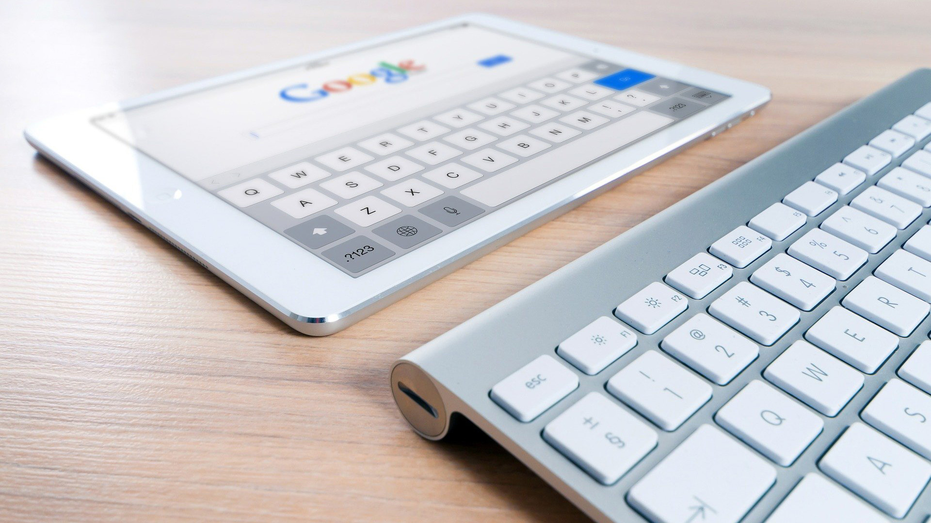 I-Pad mit Google-Logo im Display und beiliegender mobiler Tastatur liegt auf holzfarbenem Schreibtisch